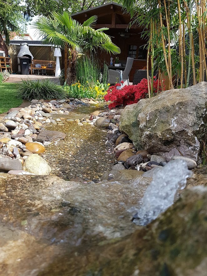 Ein kleiner Fluss in einem Privatgarten mit kleinen Steinen im Flussbett und großen Steinen am Rand. Im Hintergrund sieht man das Haus.