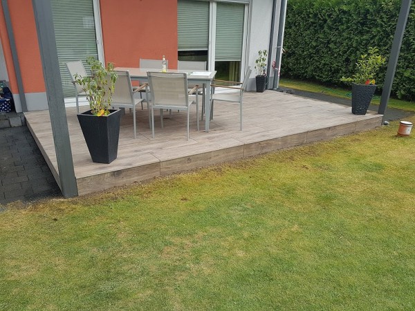 Eine Terasse aus braunen, ebenmäßigen Steinplatten. Darauf ein Tisch mit sechs Stühlen und Blumenkübeln in den Terassenecken. Vorne ist Rasenfläche und links und rechts der Terasse ist Stein verlegt.